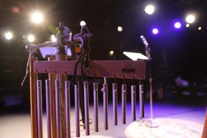 Image shows a close-up of a set of wooden and metal chimes with a background of stage lights and darkness