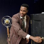 A smiling actor in brown suit opens a black prop door beside a 1940s style microphone