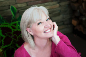 Callan, a white haired female with blue eyes looks at the camera with a big smile and a hand under her chin in a bright pink sweater.