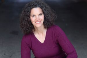 Jennifer Gatti, a female with dark curly hair and dark eyes smiles at the camera in a purple, V-neck sweater