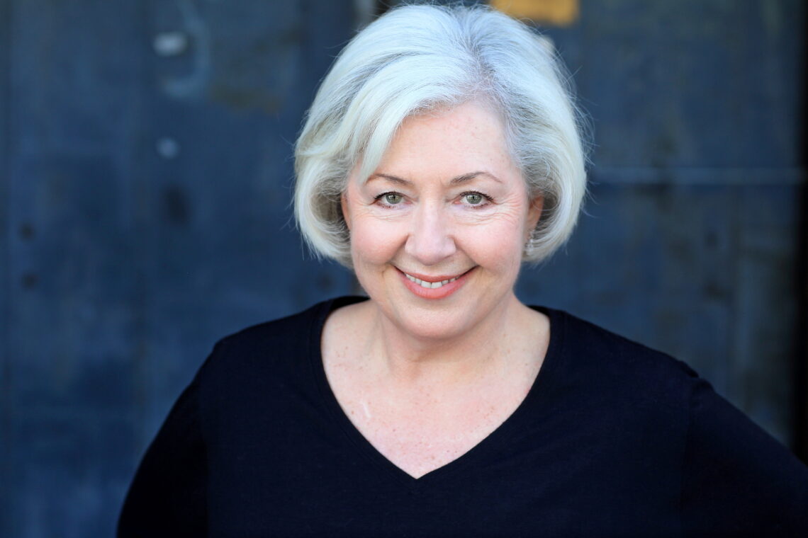 Caucasian woman with a bob of white hair and a black v-neck shirt, smiling mischievously at the camera