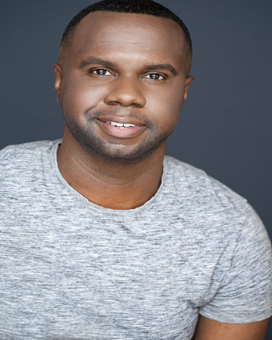 A young Black man with very short black hair and very short beard smiling at the camera and wearing a heather gray tee shirt.