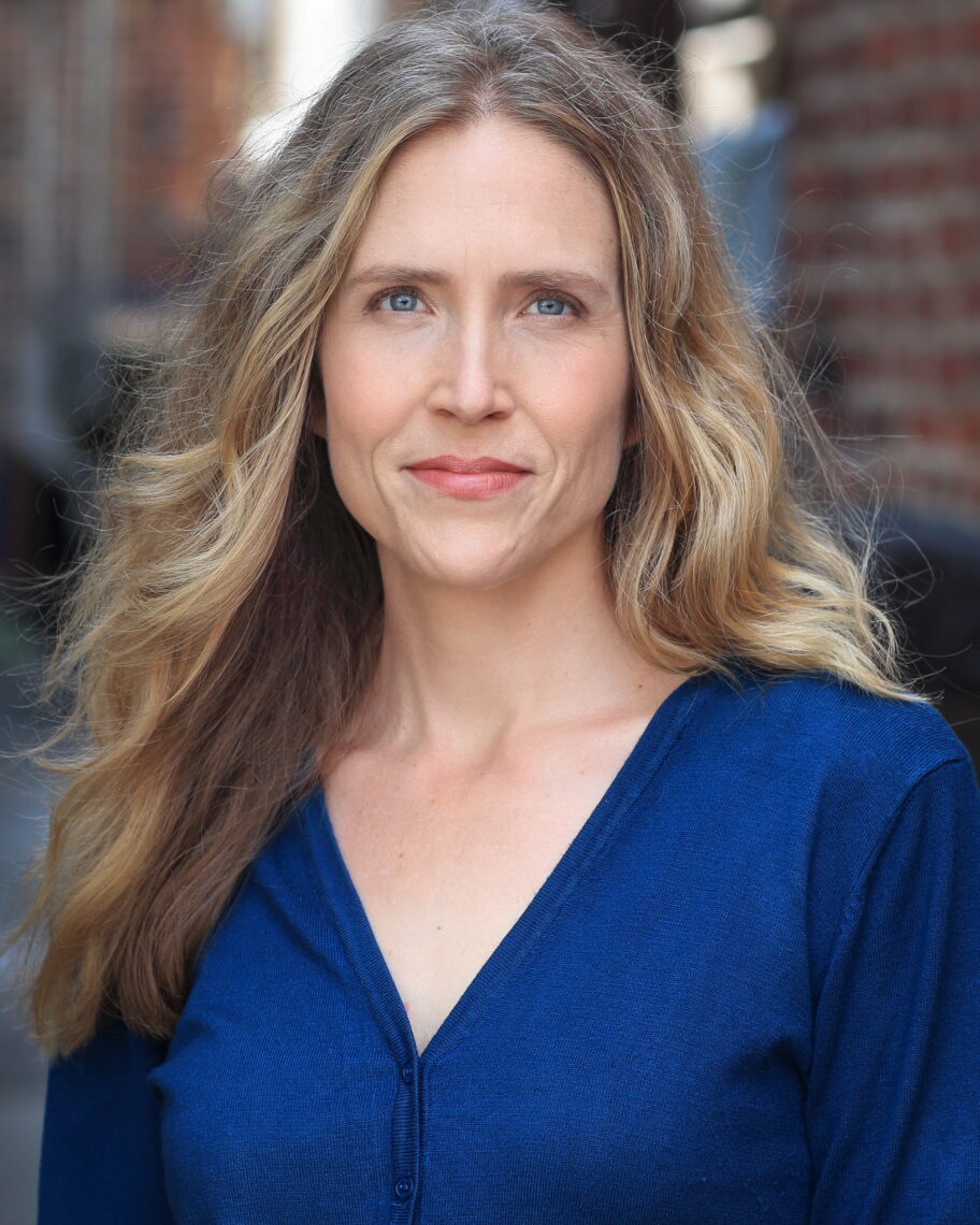 A white woman with long, wavy, blonde hair smiles without showing teeth at he camera. She is wearing a peacock blue v-neck shirt and has blue eyes.