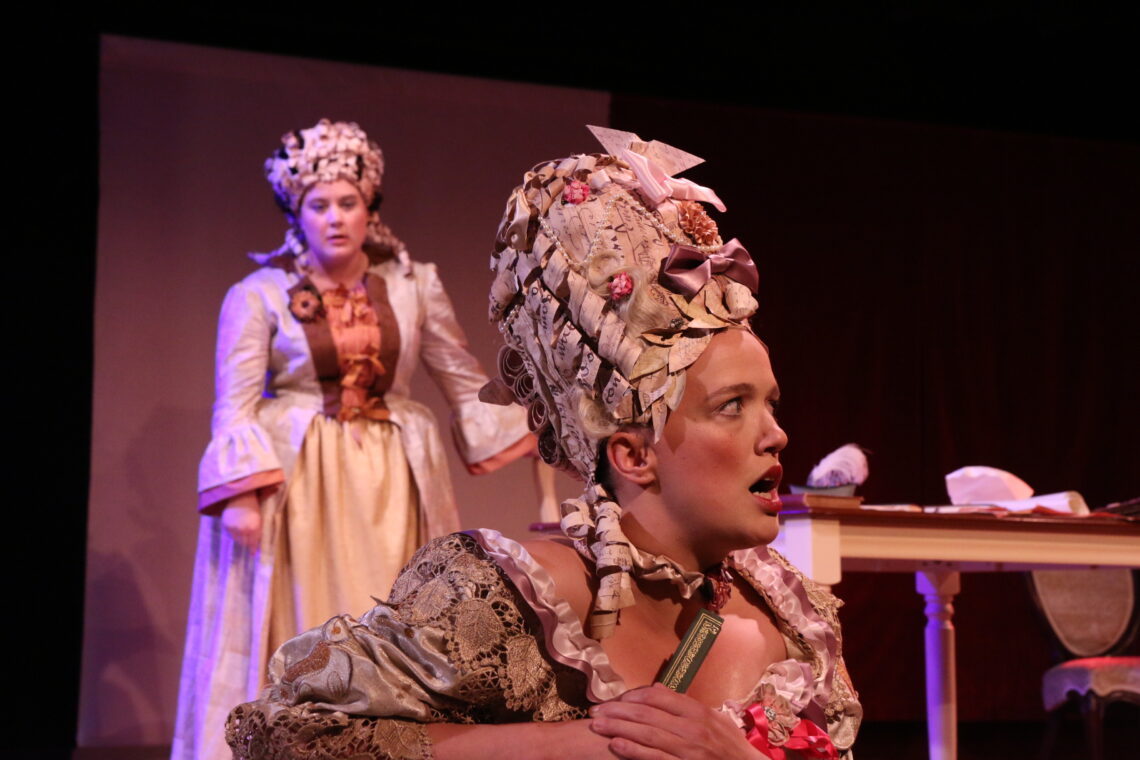 Two women in costume for the play The Revolutionists by Lauren Gunderson. The one in the foreground in dressed like Marie Antoinette in a large ribboned gown and paper wig with bows on it, is seated on the ground, and makes a surprised face, looking off camera to the right. The one in the background wears a tan colored period dress from 18th century Paris and also has a paper wig. She leans against a table and is 